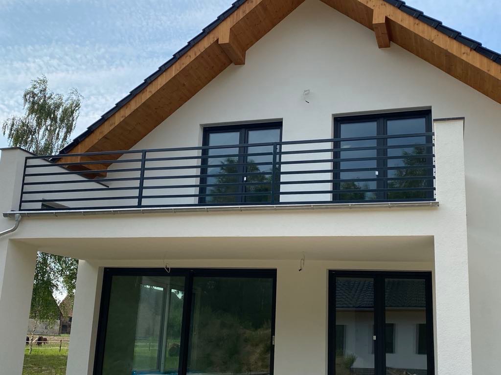 Nowoczesna, pozioma, metalowa balustrada balkonowa w kolorze antracytowym zamontowana na białym domu z drewnianym okapem dachowym. Widok na zielone drzewa odbijające się w szybach okiennych.