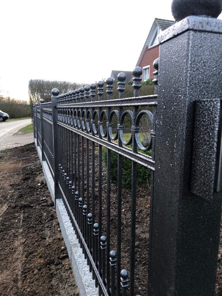 Czarne metalowe ogrodzenie z dekoracyjnymi okręgami i kulkami, zamontowane na betonowej podmurówce, widoczne z bliska pod kątem, z fragmentem domu w tle.