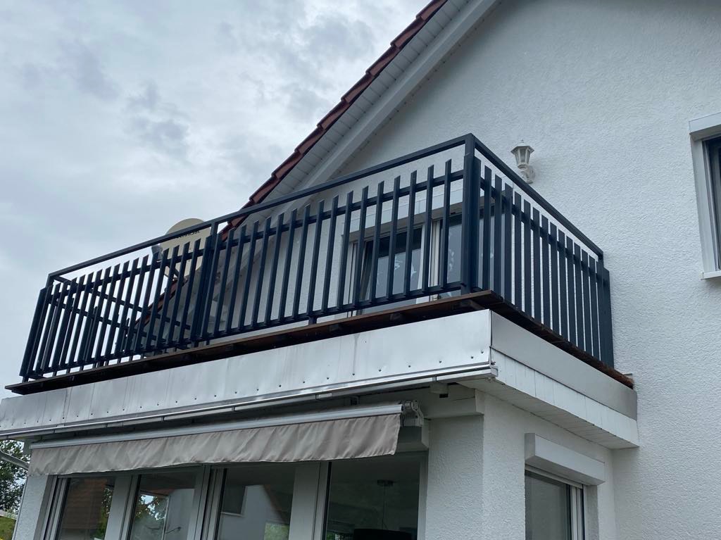 Balkon z czarną metalową balustradą o pionowych szczebelkach, zamontowany przy białym domu z widoczną anteną satelitarną i lampą ścienną.