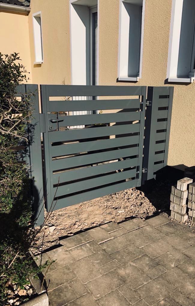 Nowoczesna, pozioma brama wjazdowa z szarych, płaskich profili, zamontowana przy jasnej elewacji budynku, widoczne przygotowane podłoże i kostka brukowa.