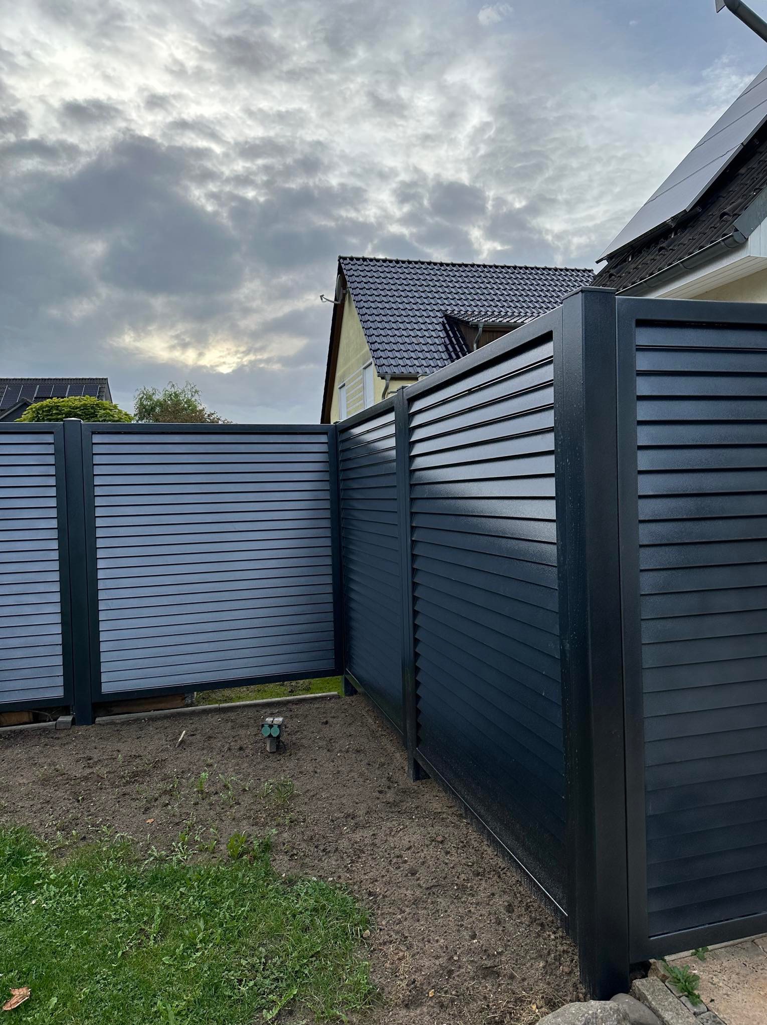 Nowoczesne, poziome, szare ogrodzenie lamelowe z metalu, widok z bliska na narożnik, z fragmentem domu w tle i pochmurnym niebem.