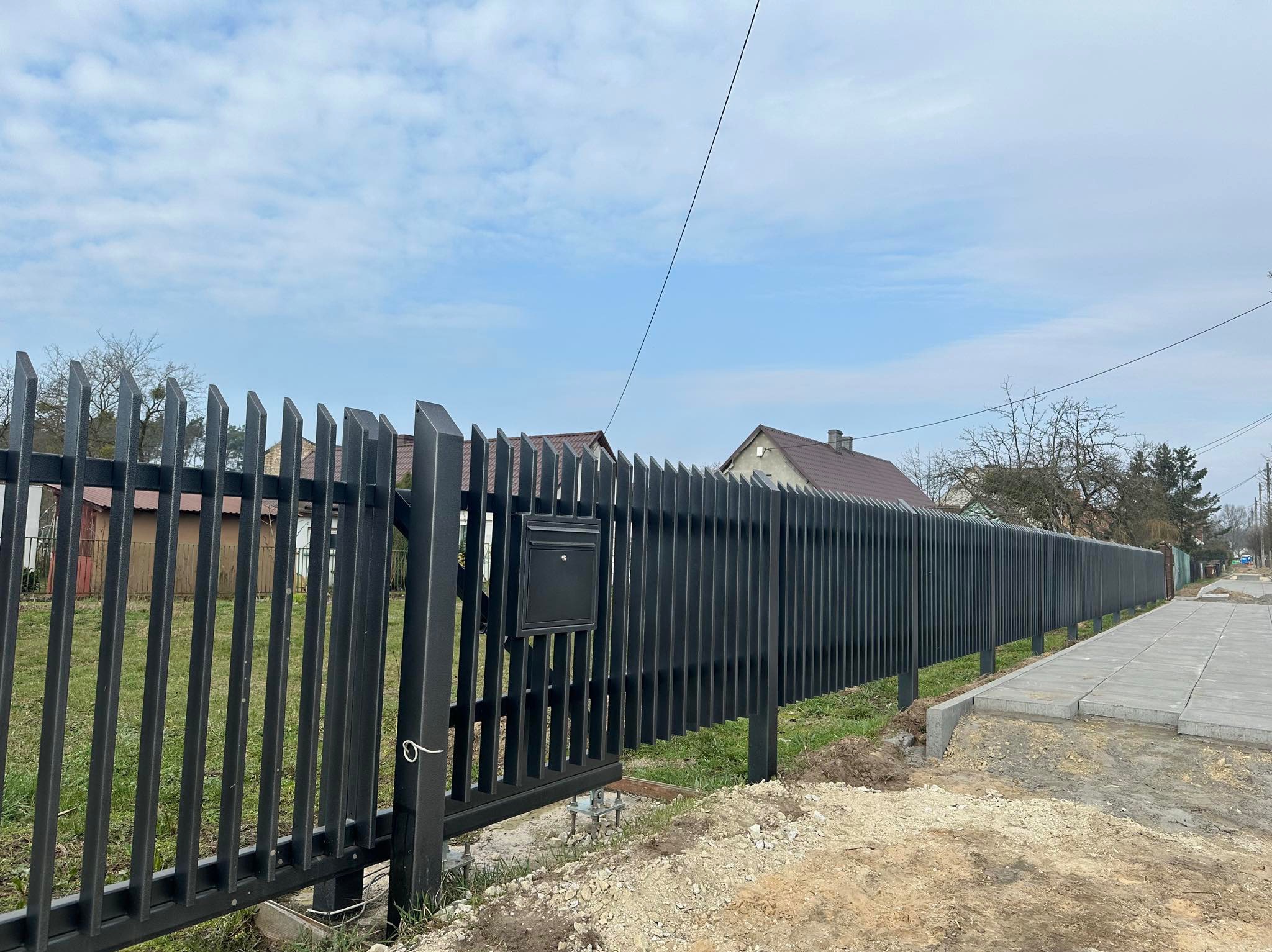 Nowoczesne, czarne ogrodzenie z pionowych, metalowych profili z bramą i skrzynką na listy, widoczne ślady prac ziemnych przy chodniku.