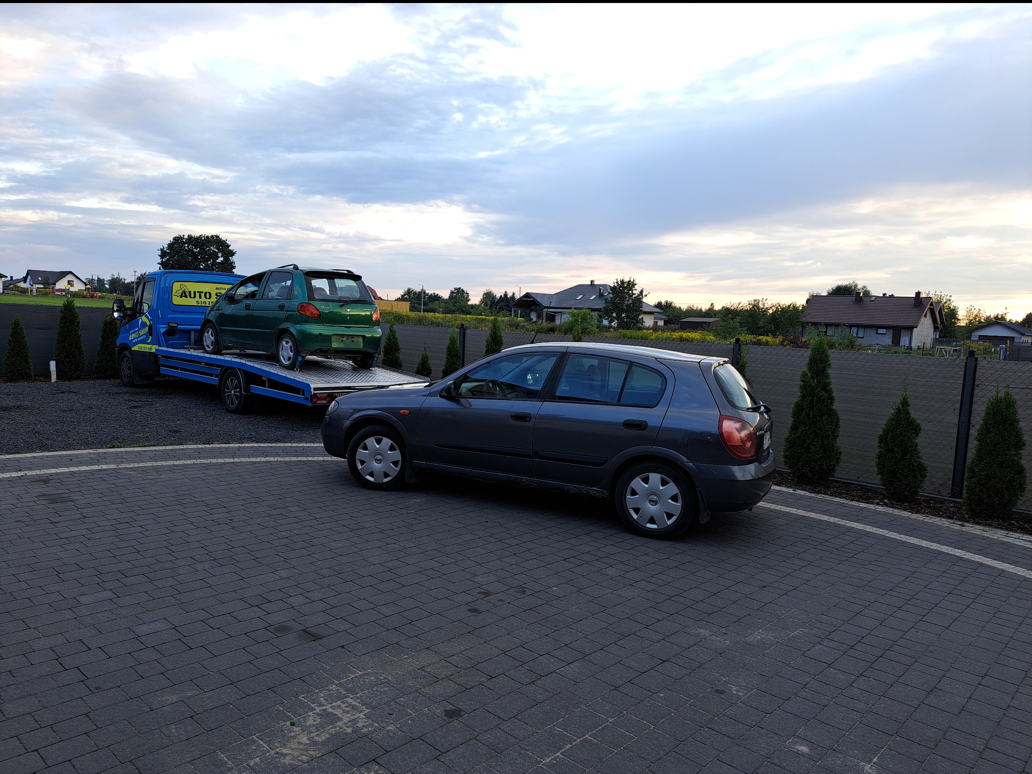 Niebieska laweta z zielonym autem na platformie, obok szary samochód osobowy na kostce brukowej, w tle zabudowania jednorodzinne i zachmurzone niebo.