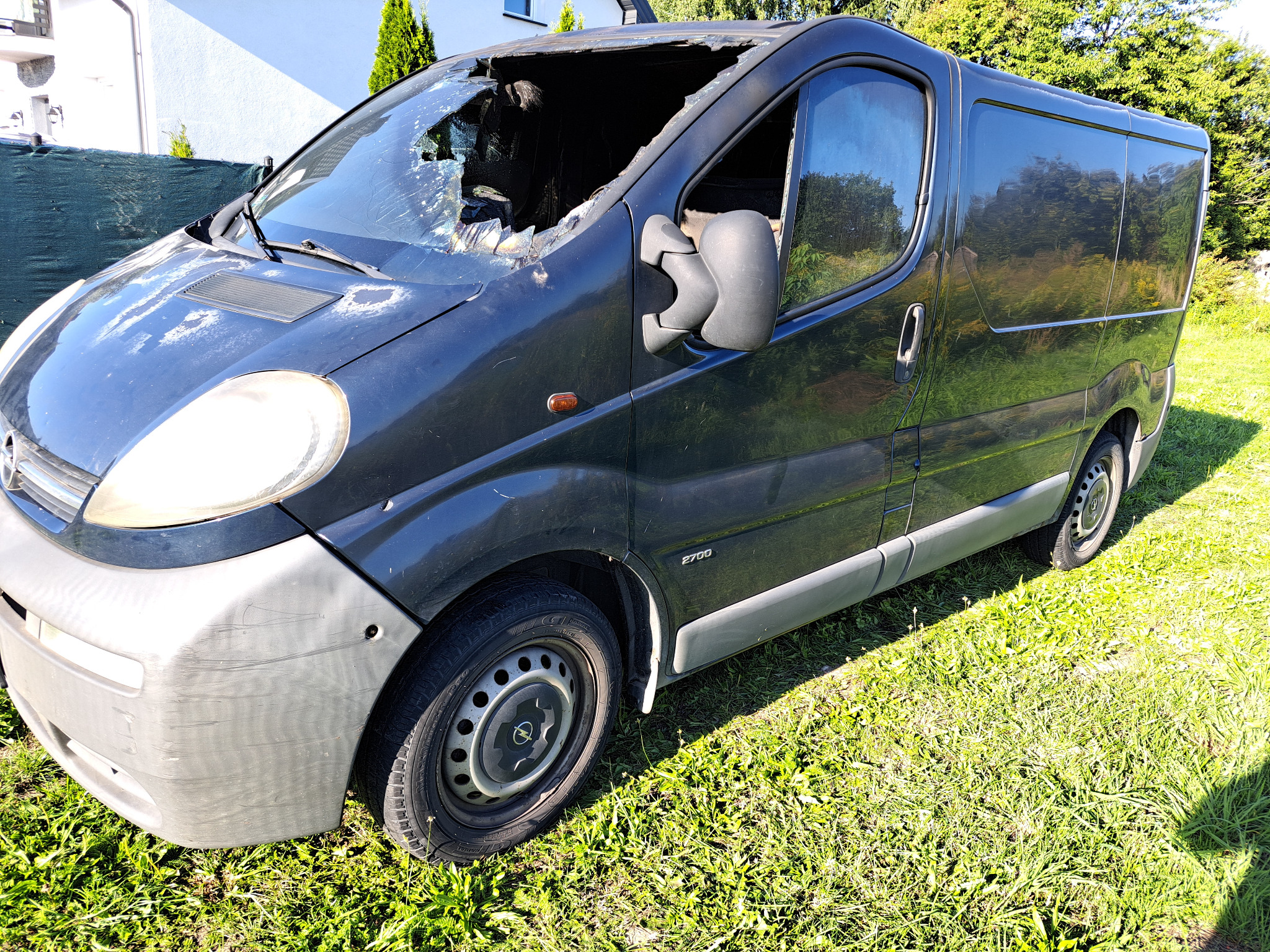 Uszkodzony, ciemny bus Opel Vivaro z powybijanymi szybami na trawie, widoczne ślady korozji i zarysowania.