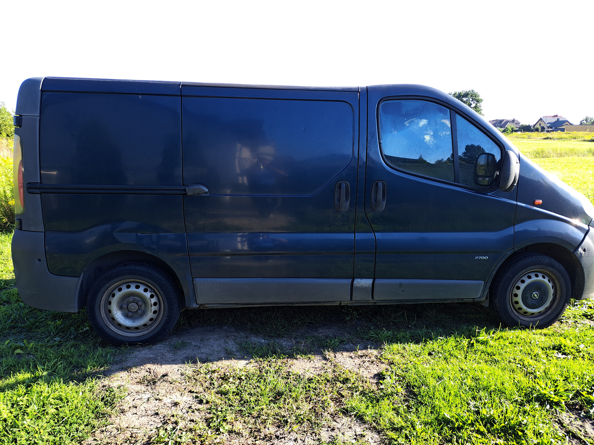 Ciemny van dostawczy z widocznym numerem 2700, zaparkowany na trawiastym terenie z widocznymi domami w tle.