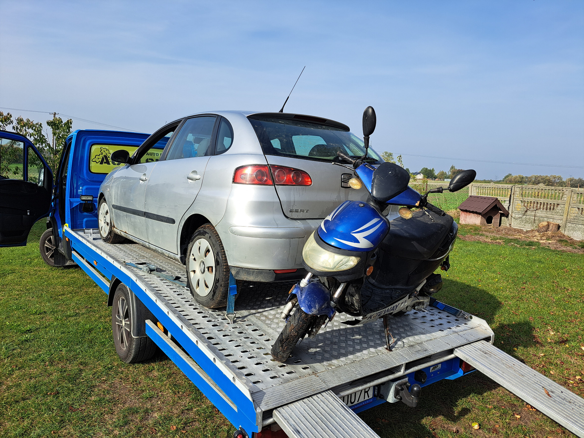 Srebrny Seat Ibiza i niebieski skuter Keeway na platformie lawety Auto Złom z otwartymi drzwiami kabiny, na tle zielonej trawy i błękitnego nieba.