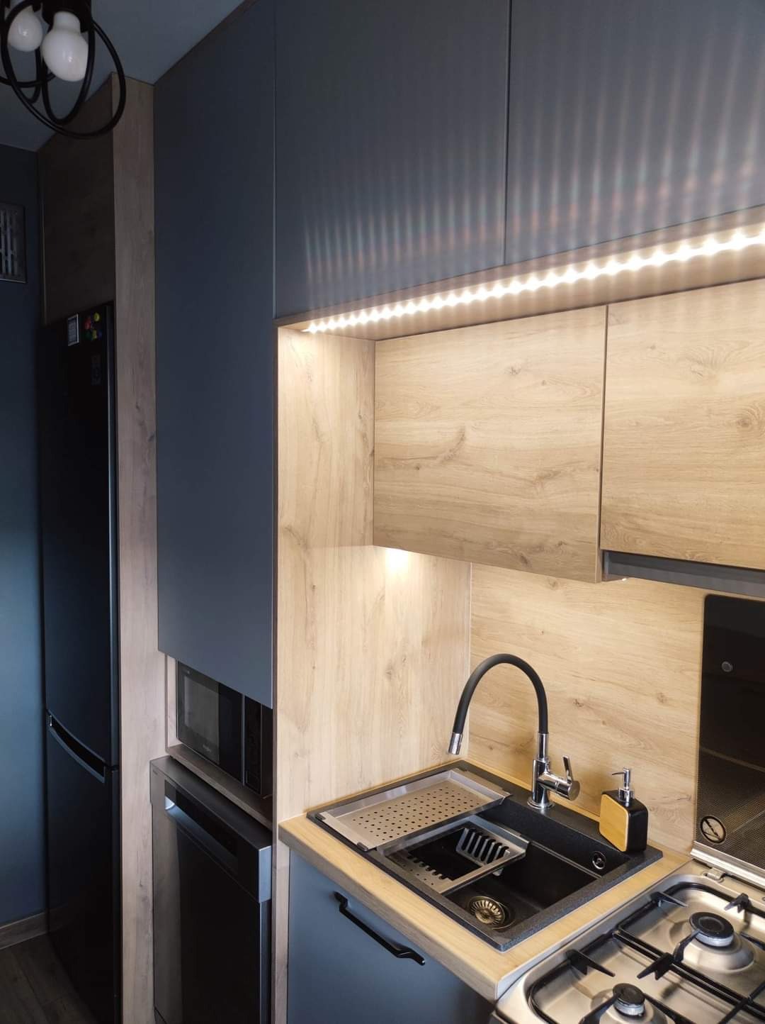 Nowoczesna kuchnia z szarymi szafkami, drewnopodobnym wykończeniem, podświetleniem LED, czarnym zlewem z ociekaczem i czarną baterią, oraz czarną lodówką side-by-side.