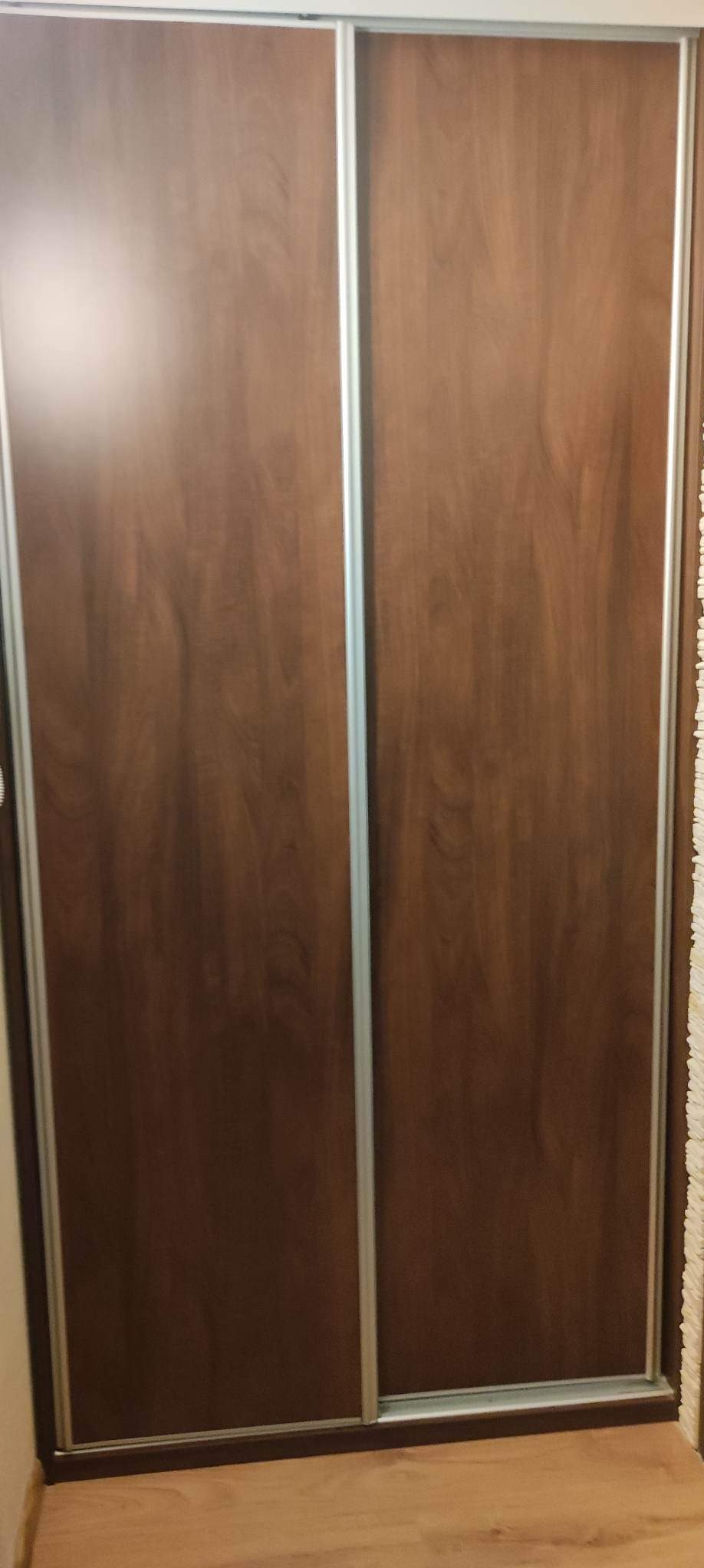 Brązowa szafa przesuwna z drewnopodobnymi frontami i srebrnymi prowadnicami, widoczna z bliska na tle podłogi.