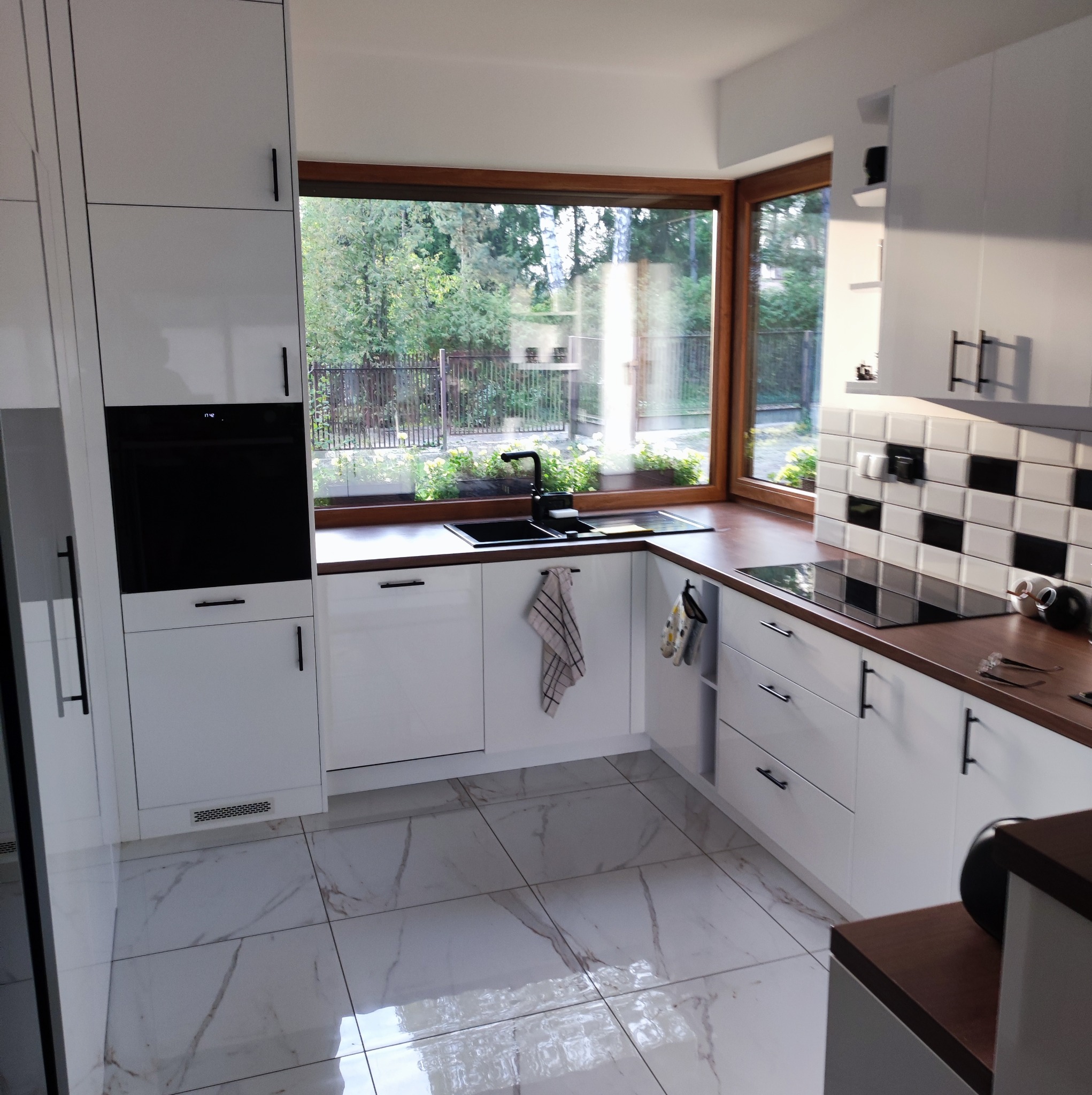 Nowoczesna, jasna kuchnia z białymi szafkami, drewnianym blatem i czarnymi akcentami, widok na ogród przez duże okno.