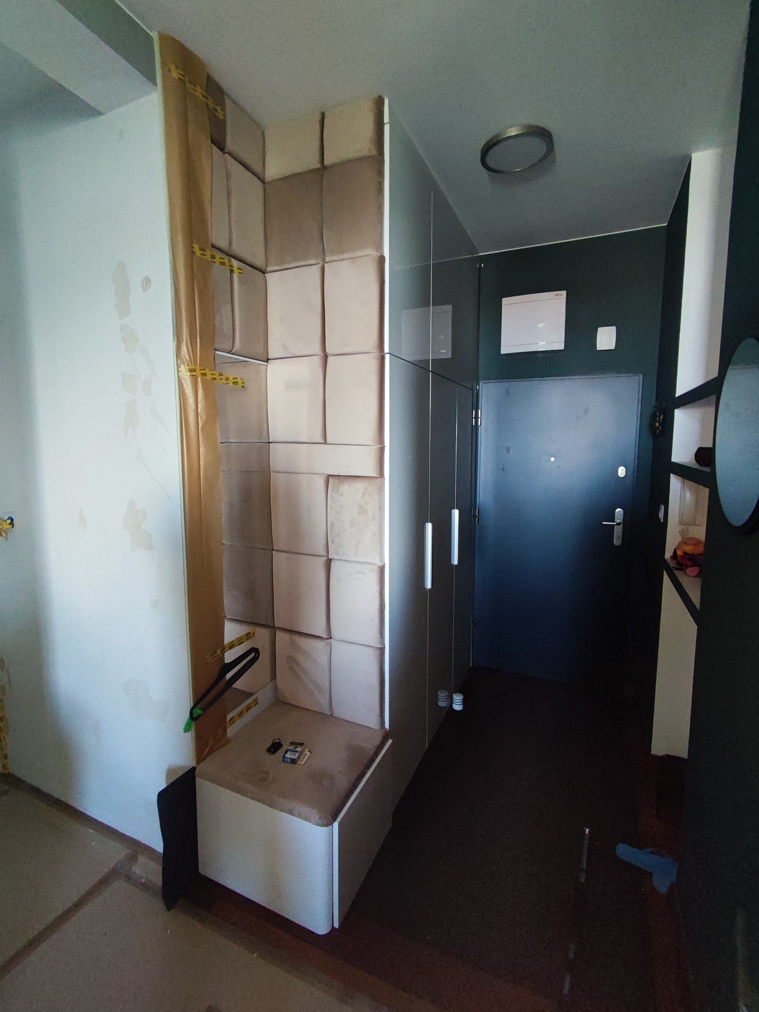 Fragment przedpokoju w trakcie realizacji zabudowy z tapicerowanych paneli ściennych, widoczne zabezpieczenia folią malarską, obok szafa wnękowa z białymi frontami.