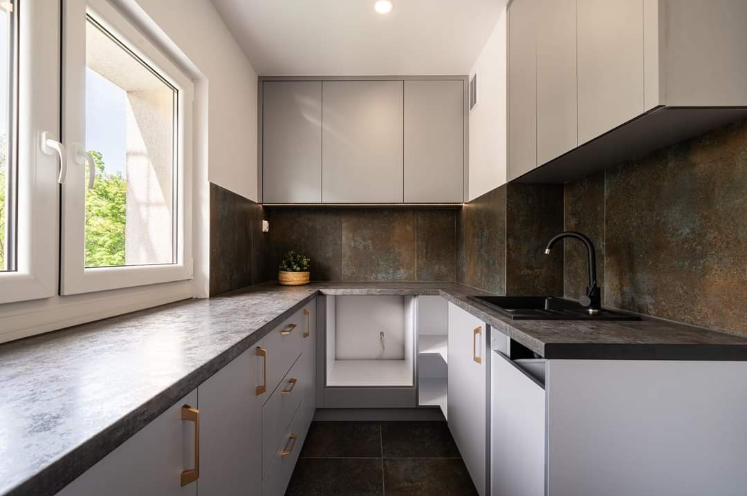 Nowoczesna kuchnia w kształcie litery U z szarymi szafkami, blatem imitującym kamień, czarnym zlewem i baterią, oraz dekoracyjną ścianą nad blatem, oświetlona punktowo.