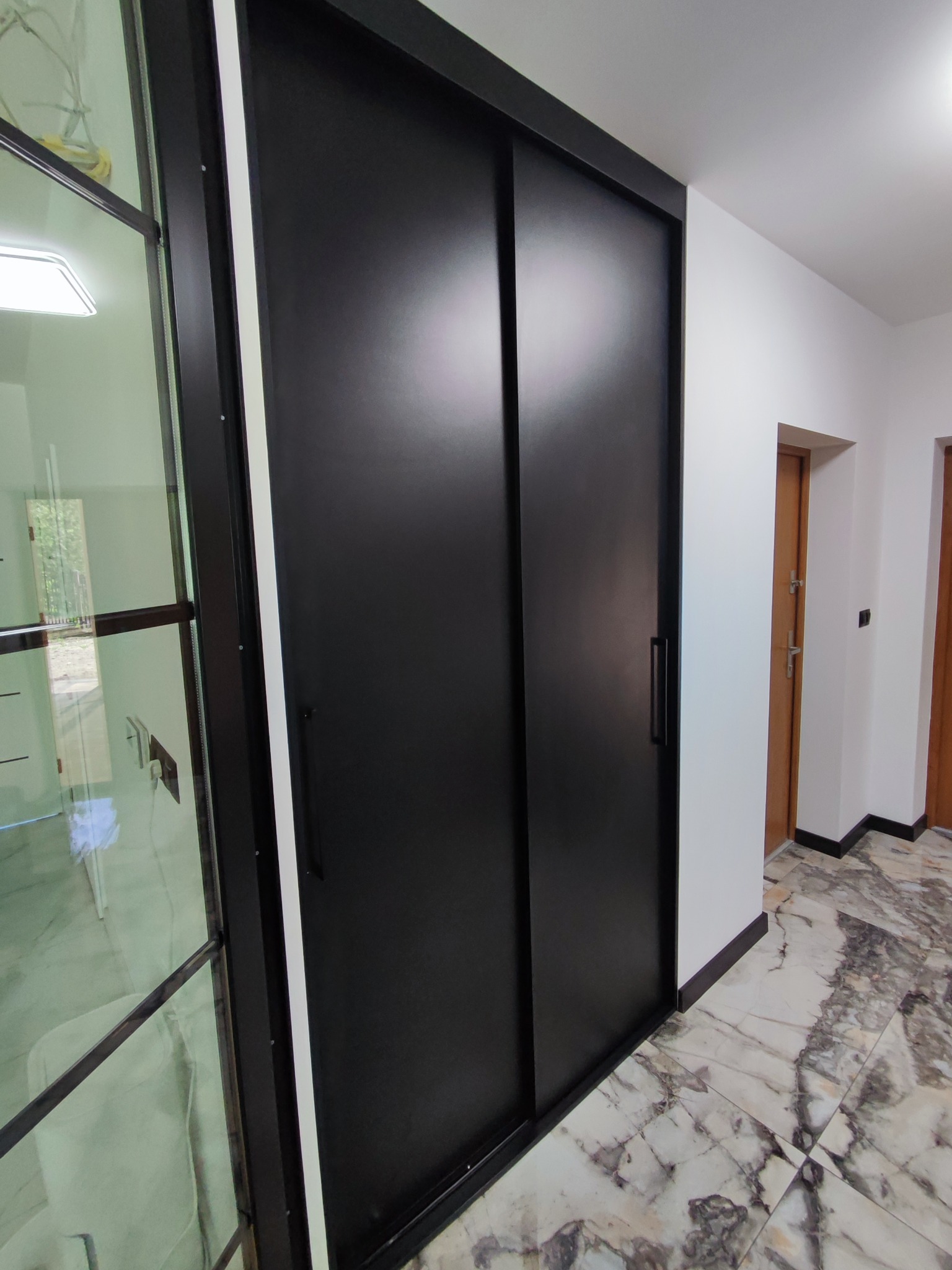 Czarna szafa przesuwna z matowymi drzwiami i minimalistycznymi uchwytami, widoczna z perspektywy podłogi z marmurowym wzorem, obok przeszklonej ściany działowej.