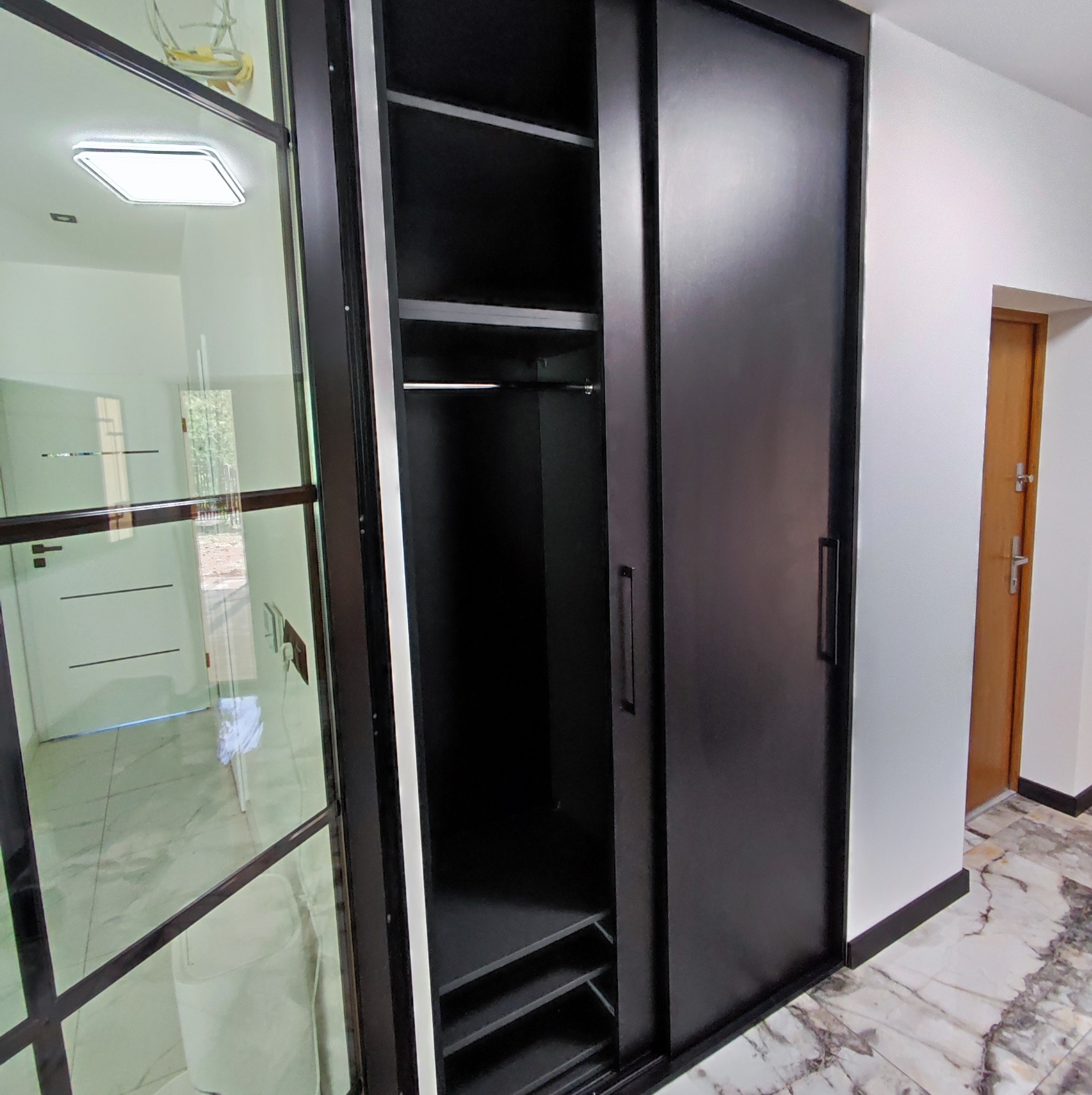 Czarna szafa wnękowa z przesuwnymi drzwiami, widoczne półki i drążek na ubrania, marmurowa podłoga, drzwi wejściowe w tle.