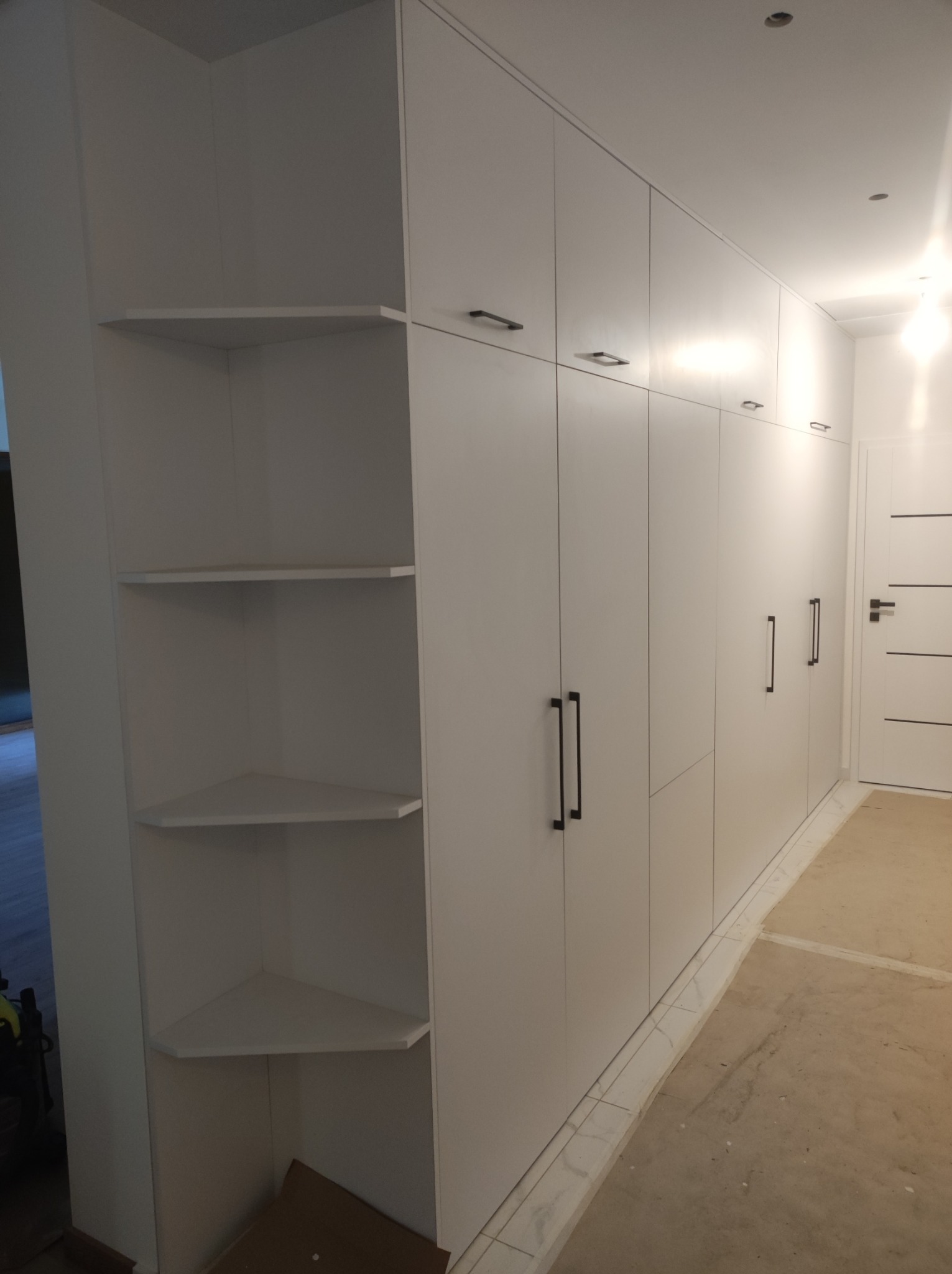 Biała szafa wnękowa z półkami narożnymi i długimi czarnymi uchwytami, widok na korytarz z białymi drzwiami.