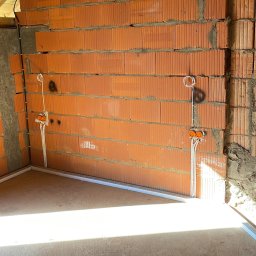 MK Elektro - Surowy mur z pustaków ceramicznych z rozprowadzoną instalacją elektryczną: kable w peszlach prowadzone pionowo od puszek instalacyjnych, widoczne elementy konstrukcji dachu