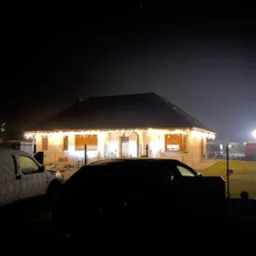 Nocne ujęcie domu jednorodzinnego z dekoracyjnym oświetleniem zewnętrznym, częściowo zasłoniętego przez zaparkowane pojazdy.