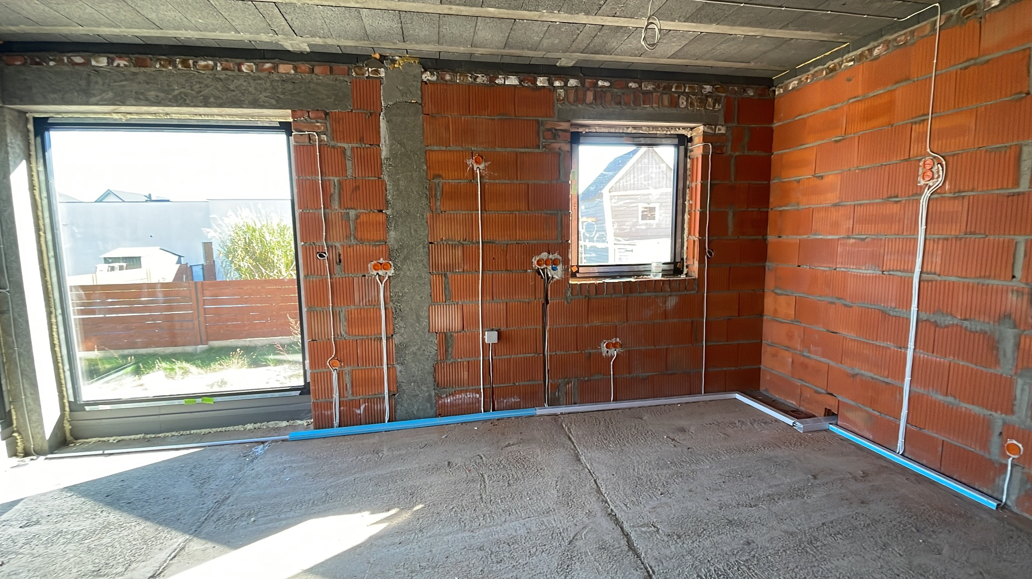 Surowy stan wnętrza z widocznymi przewodami elektrycznymi i puszkami instalacyjnymi na ścianach z cegły dziurawki, okna wstawione, brak tynków, wylewka na podłodze, listwy przypodłogowe częściowo...