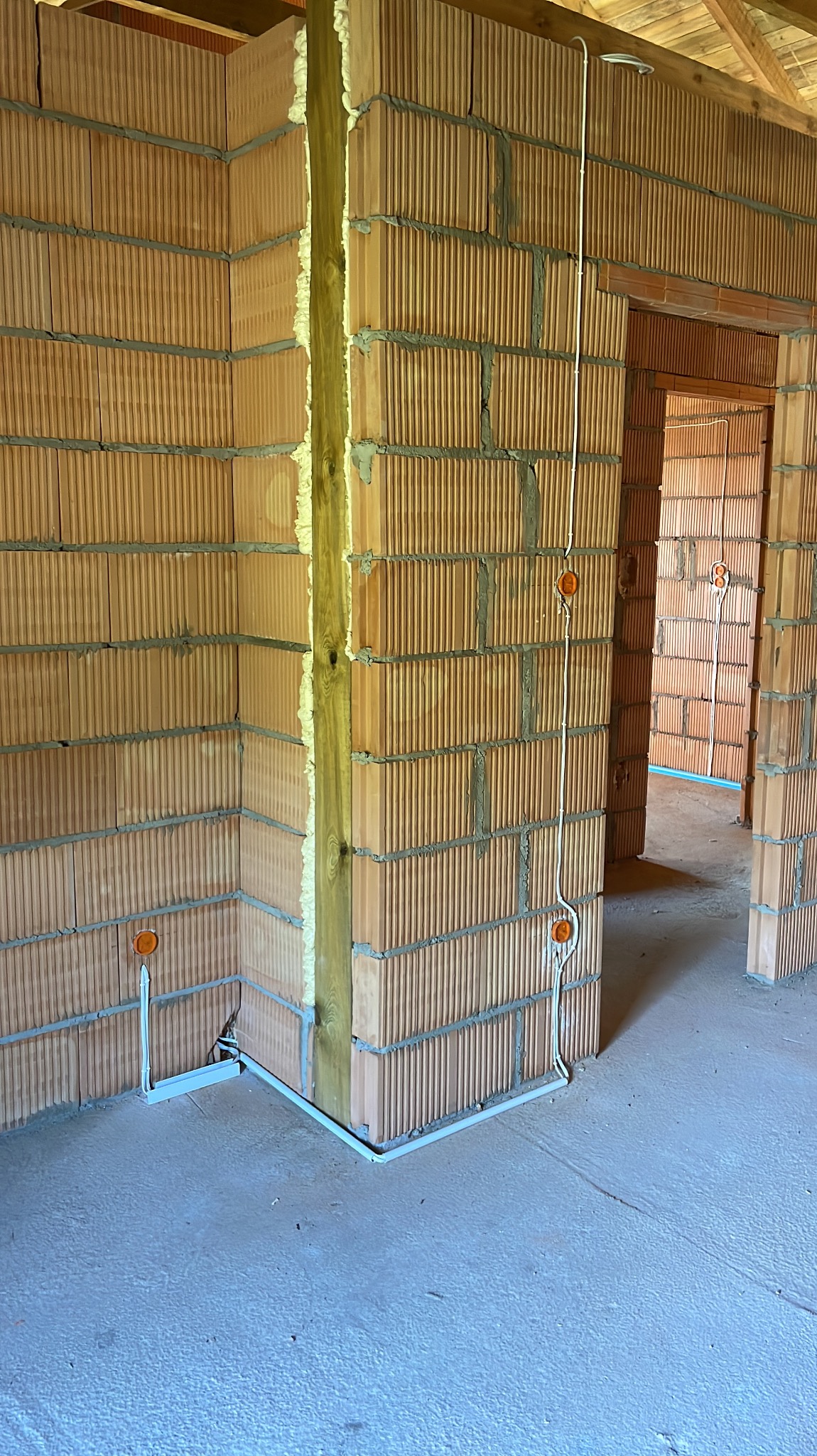 Wnętrze w trakcie budowy: ściany z cegły dziurawki, pionowy słup drewniany z pianką montażową, białe rurki instalacji elektrycznej z pomarańczowymi puszkami podtynkowymi, surowa podłoga.