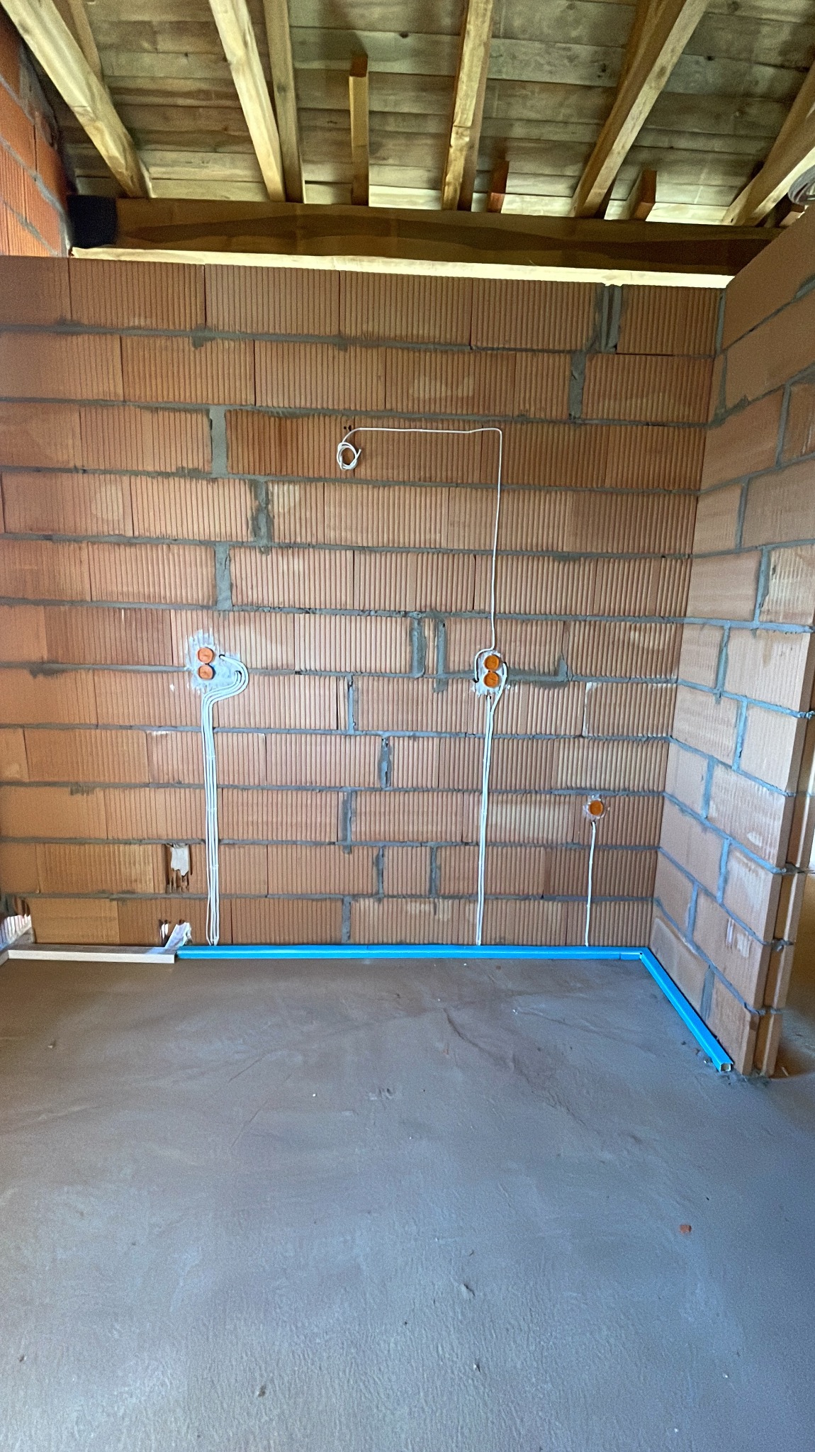 Surowy mur z cegły dziurawki z poprowadzonymi przewodami elektrycznymi i puszkami pod gniazdka, widoczne także niebieskie rurki instalacyjne przy podłodze i drewniany strop.