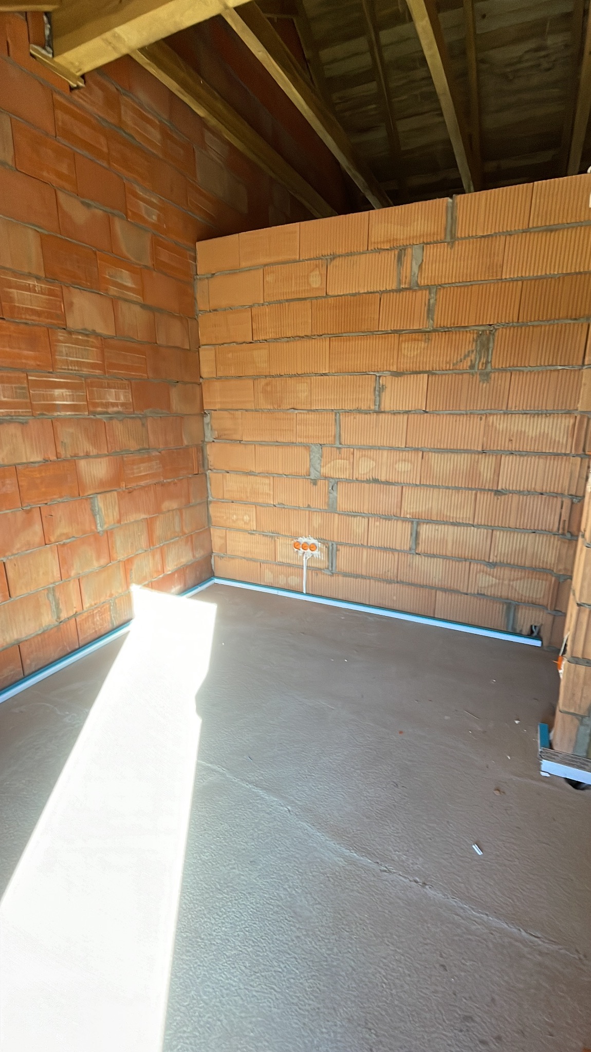 Surowy stan wnętrza z ceglanymi ścianami i wylewką podłogową, widoczne puszki elektryczne w ścianie, przygotowane do dalszych prac instalacyjnych.
