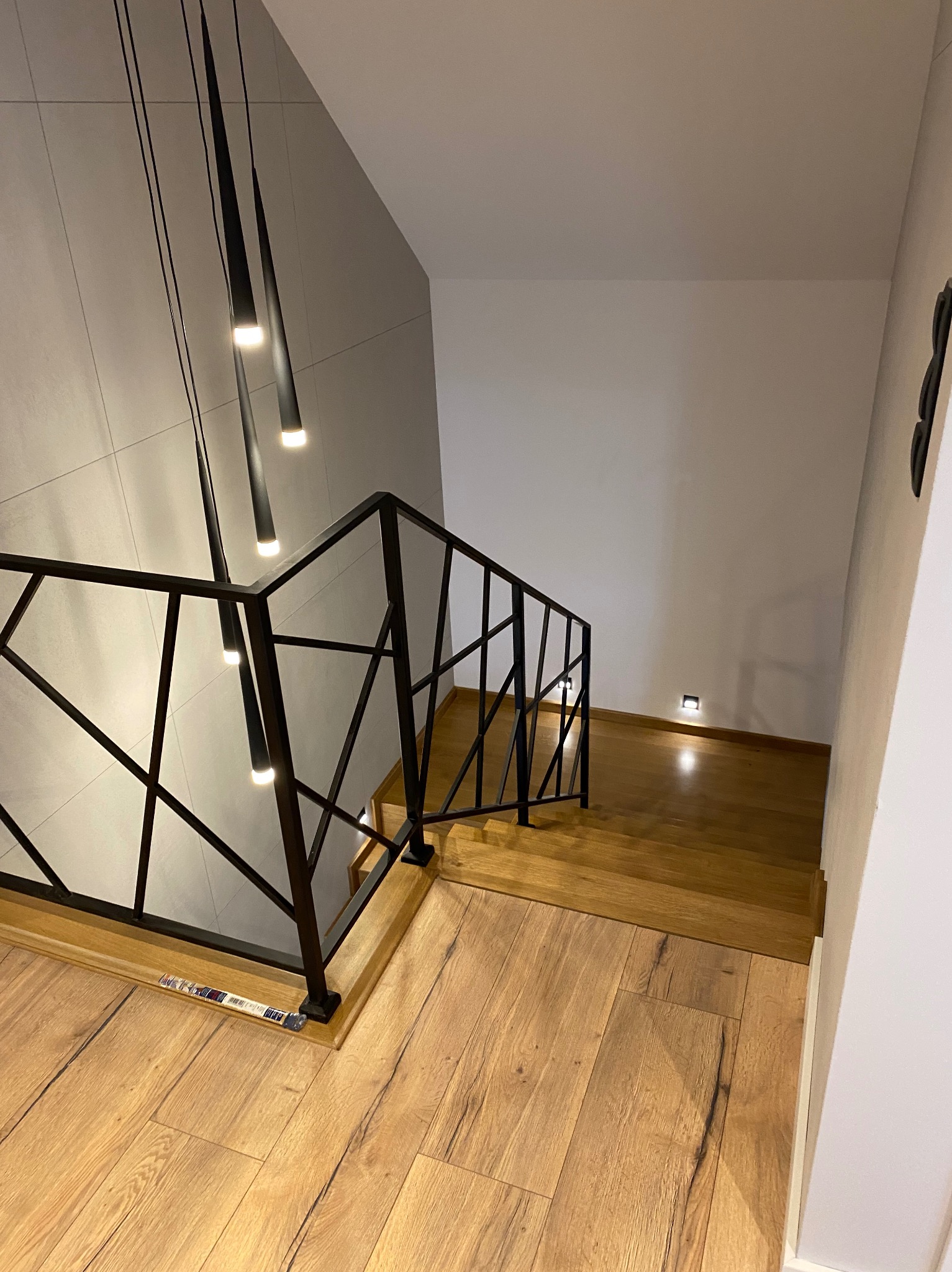Nowoczesne oświetlenie schodów z czarnymi, wiszącymi lampami i podświetleniem stopni, widok z góry na drewniane schody z czarną, geometryczną balustradą.