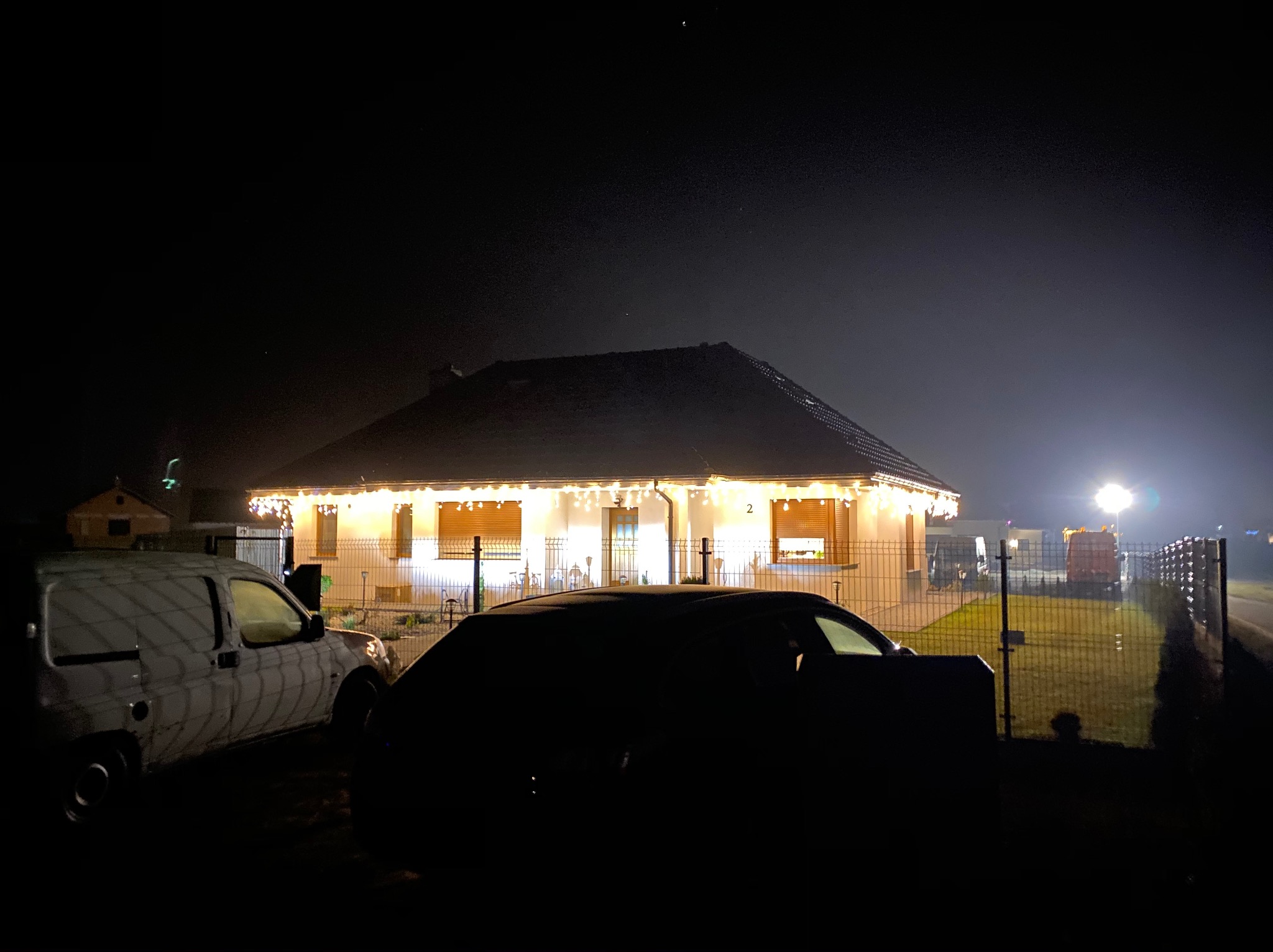 Nocne ujęcie domu jednorodzinnego z dekoracyjnym oświetleniem zewnętrznym, częściowo zasłoniętego przez zaparkowane pojazdy.