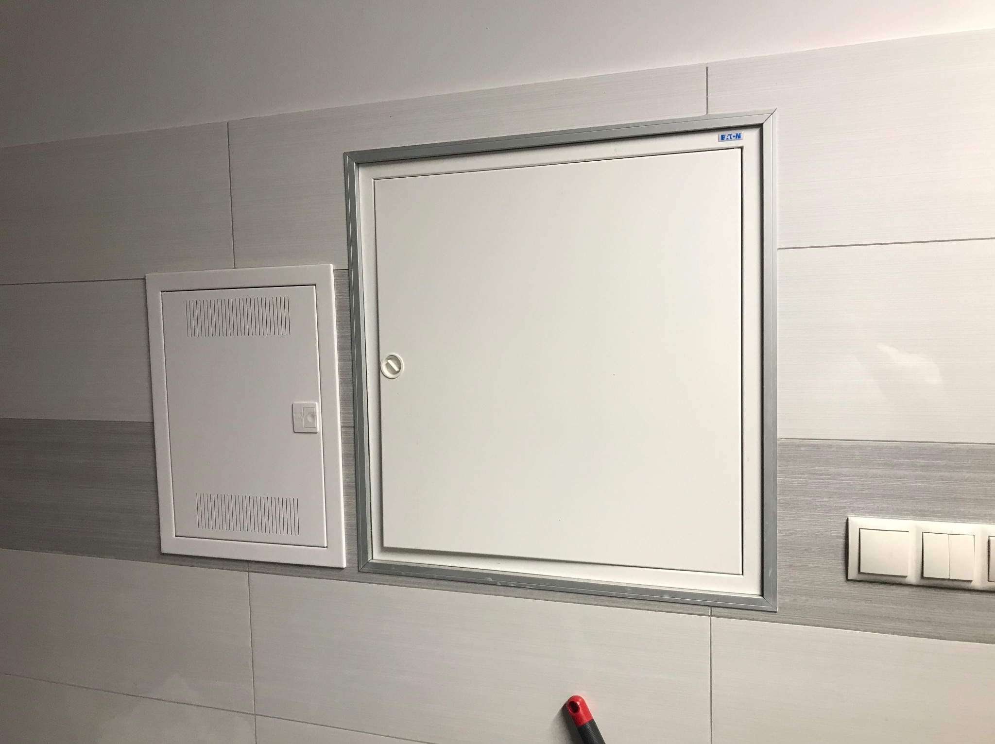 Biała skrzynka elektryczna z kratką wentylacyjną i kwadratowa klapa rewizyjna z metalową ramką, umieszczone na ścianie wyłożonej płytkami w odcieniach szarości, obok włączniki światła.