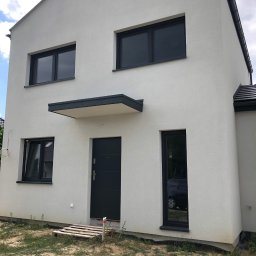 Pmbudex - Nowoczesny, dwukondygnacyjny dom z elewacją w jasnym odcieniu szarości, antracytowymi oknami i drzwiami, widoczny z zewnątrz w słoneczny dzień, z paletą leżącą na ziemi przy wejściu.