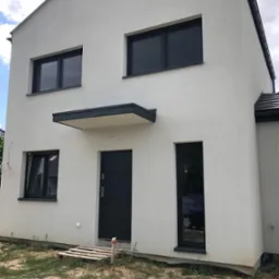 Nowoczesny, dwukondygnacyjny dom z elewacją w jasnym odcieniu szarości, antracytowymi oknami i drzwiami, widoczny z zewnątrz w słoneczny dzień, z paletą leżącą na ziemi przy wejściu.