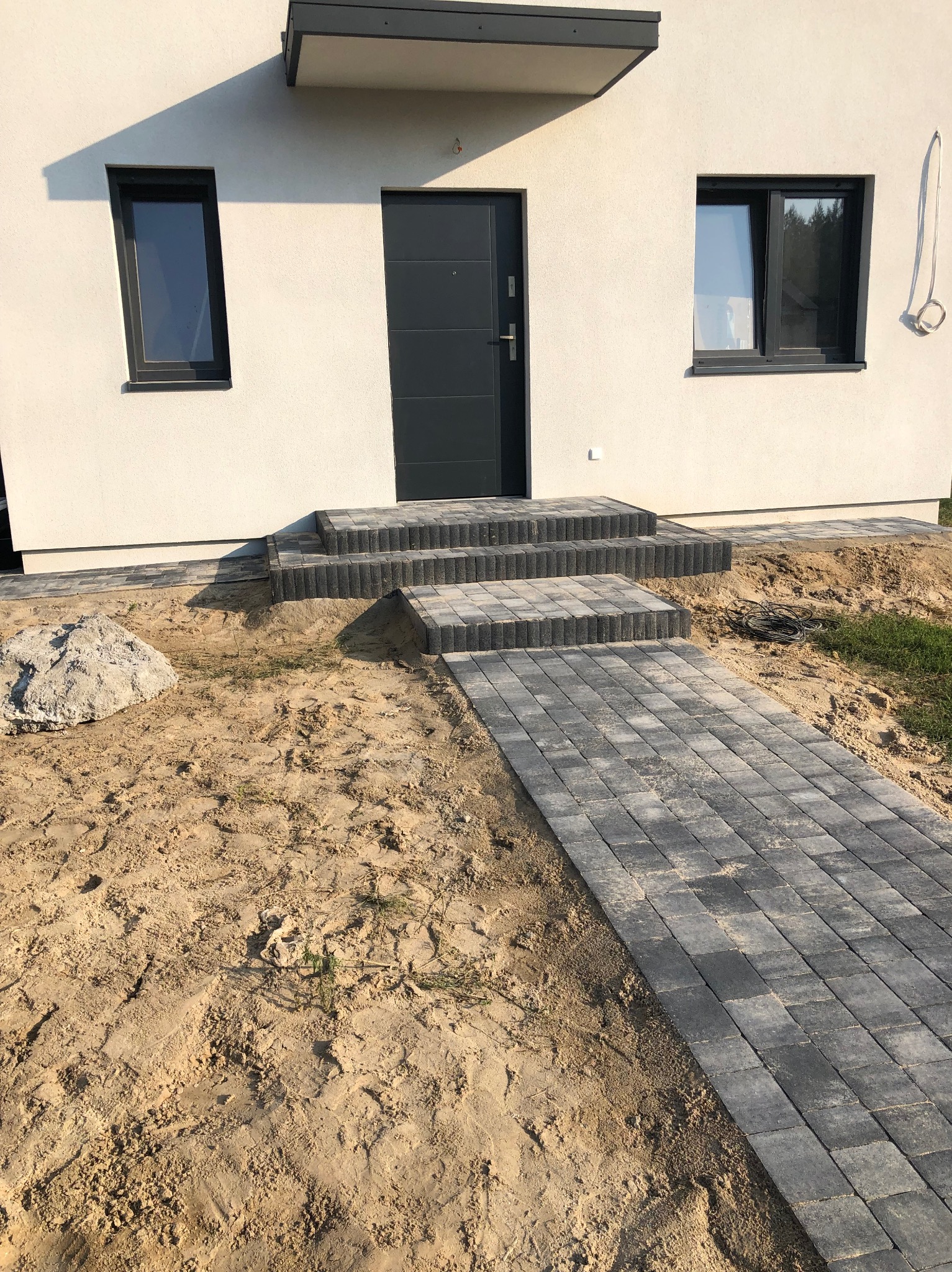 Nowoczesny dom jednorodzinny z szarą stolarką okienną i drzwiową oraz brukowanym wejściem i ścieżką, teren wokół w trakcie prac ziemnych.
