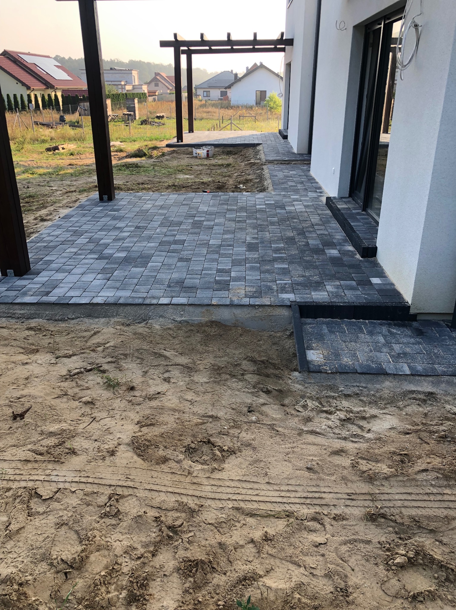 Układanie kostki brukowej w kolorze szarym, tworzącej ścieżkę i schody przy nowoczesnym domu z białymi ścianami. Widoczny fragment drewnianej pergoli i przygotowane podłoże z piasku.