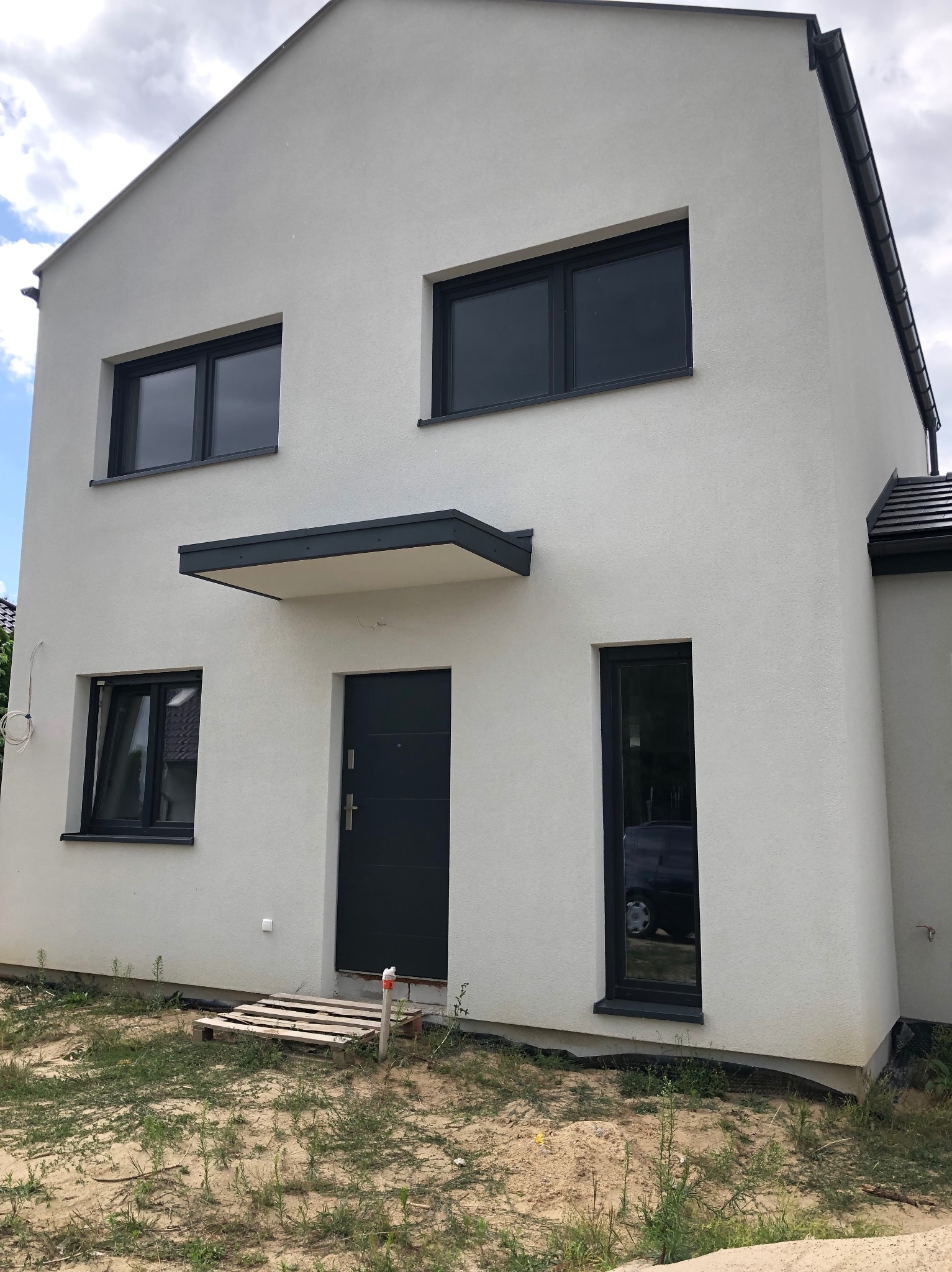 Nowoczesny, dwukondygnacyjny dom z elewacją w jasnym odcieniu szarości, antracytowymi oknami i drzwiami, widoczny z zewnątrz w słoneczny dzień, z paletą leżącą na ziemi przy wejściu.