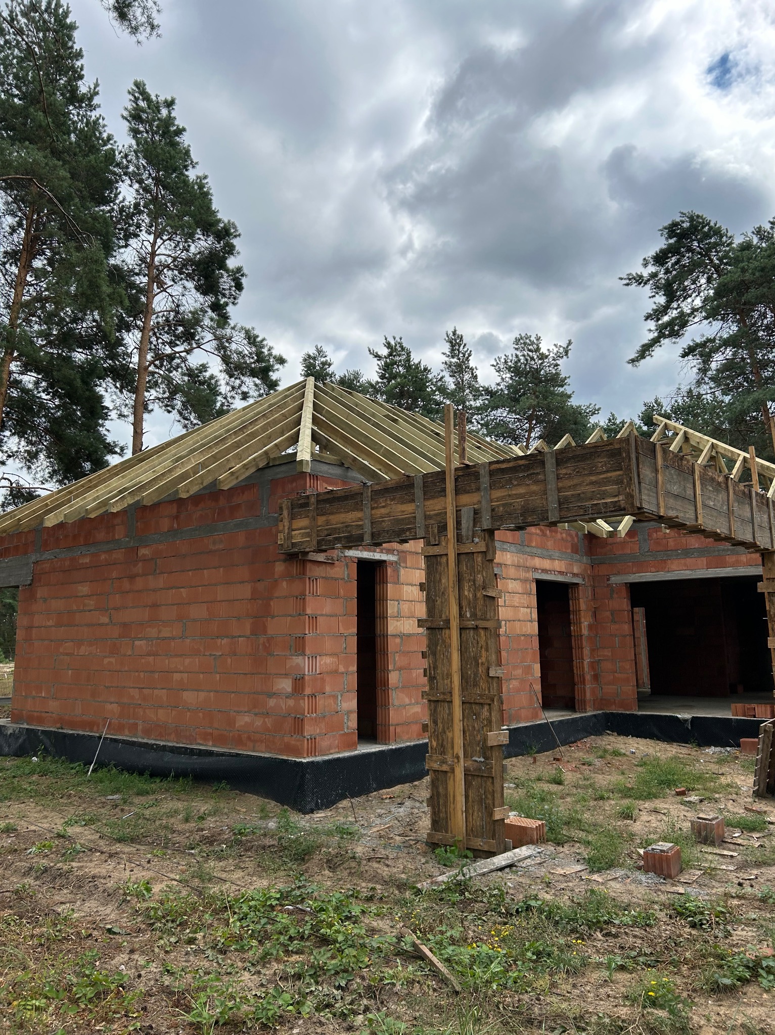 Surowy stan budowy domu jednorodzinnego z czerwonej cegły, widoczna konstrukcja dachu z drewnianych belek i szalunek nad otworami okiennymi, fundament zabezpieczony czarną folią izolacyjną, w tle...