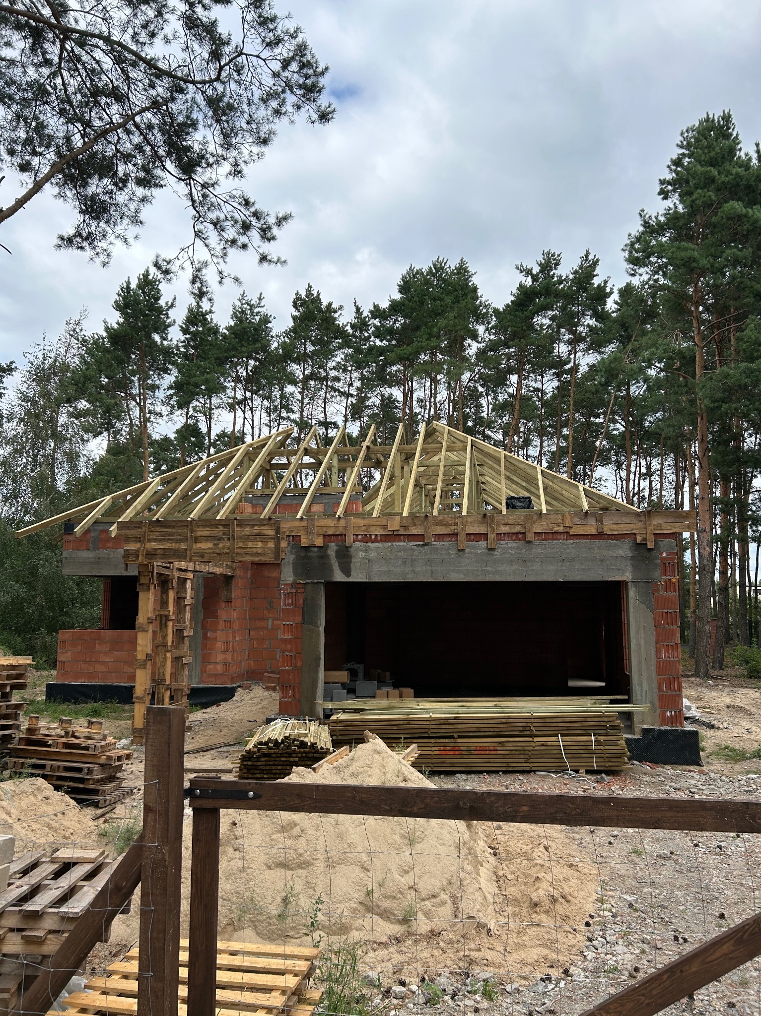 Surowy szkielet dachu z drewnianych belek na murowanym budynku w stanie surowym otwartym, widoczne palety i materiały budowlane na posesji.