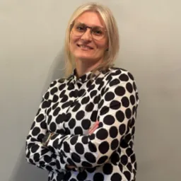 Portret uśmiechniętej blondynki w okularach i koszuli w grochy, z założonymi rękami na tle szarej ściany.