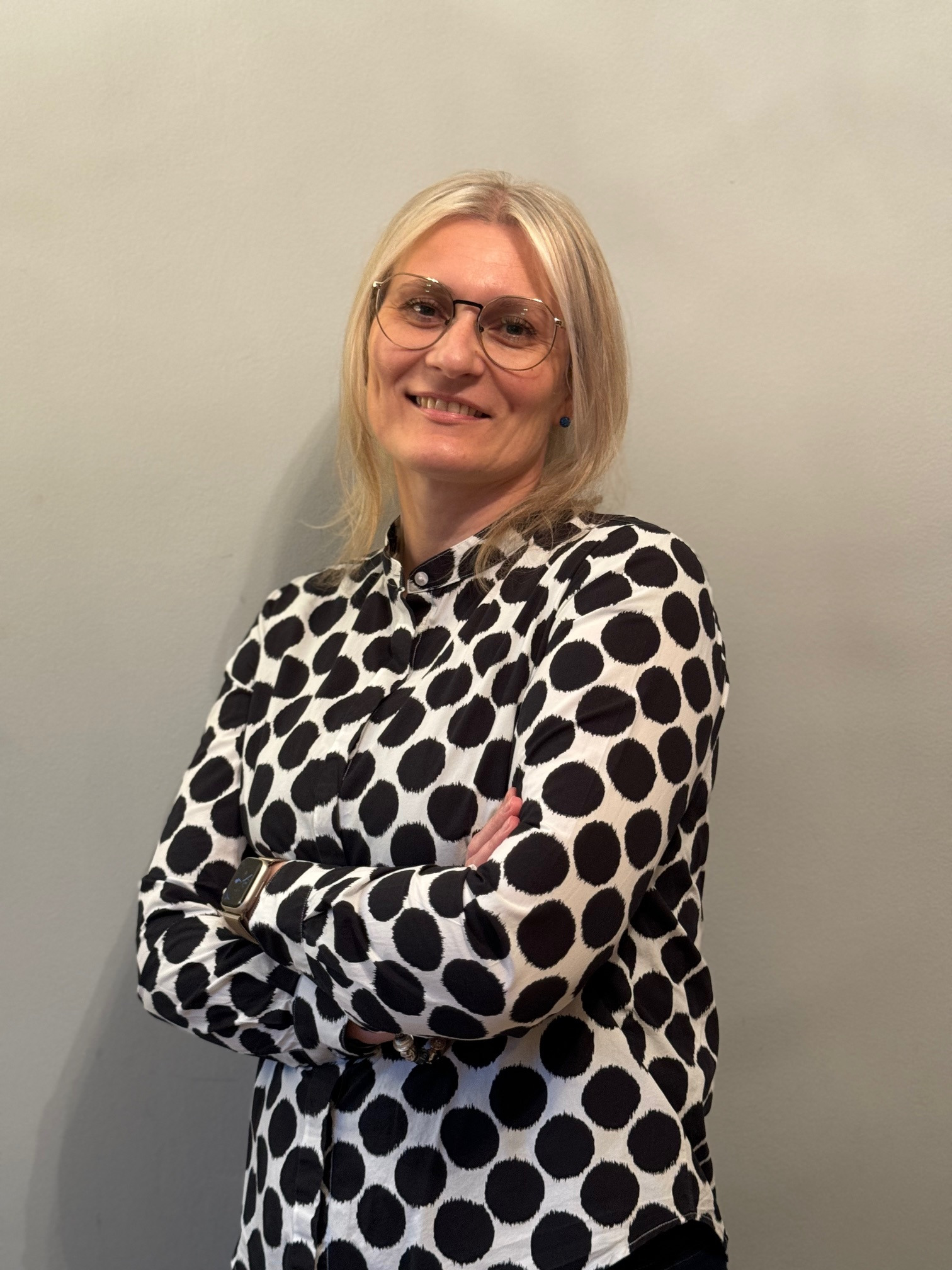 Portret uśmiechniętej blondynki w okularach i koszuli w grochy, z założonymi rękami na tle szarej ściany.