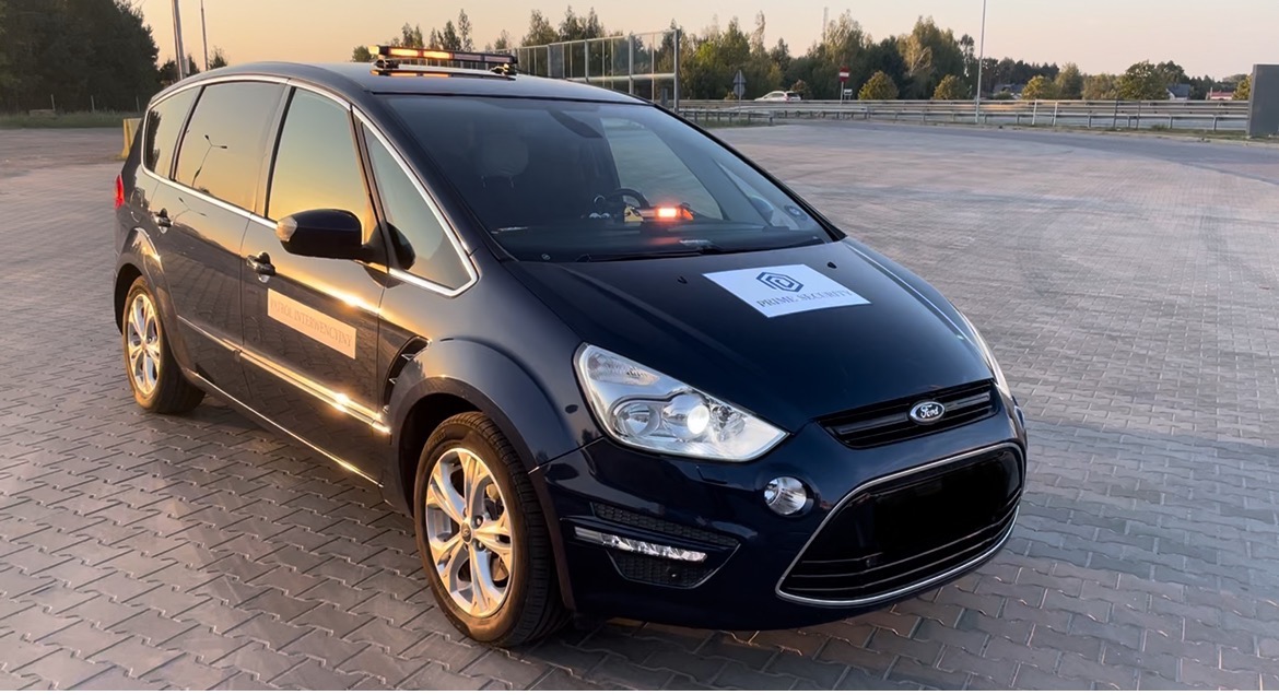 Ciemnoniebieski samochód Ford z oznaczeniami 'PRIME SECURITY' i napisem 'POJAZD INTERWENCYJNY' na parkingu z kostki brukowej, wyposażony w sygnalizację świetlną na dachu i desce rozdzielczej.