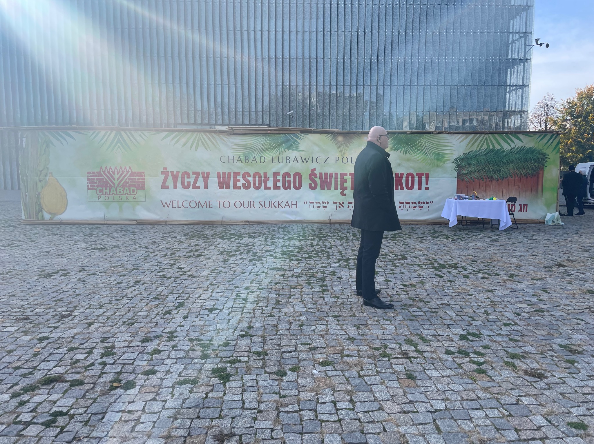 Konstrukcja Sukkah z banerem 'Życzy Wesołego Święta Sukkot' na tle nowoczesnego budynku ze szklaną fasadą, mężczyzna w czarnym płaszczu stoi z boku.