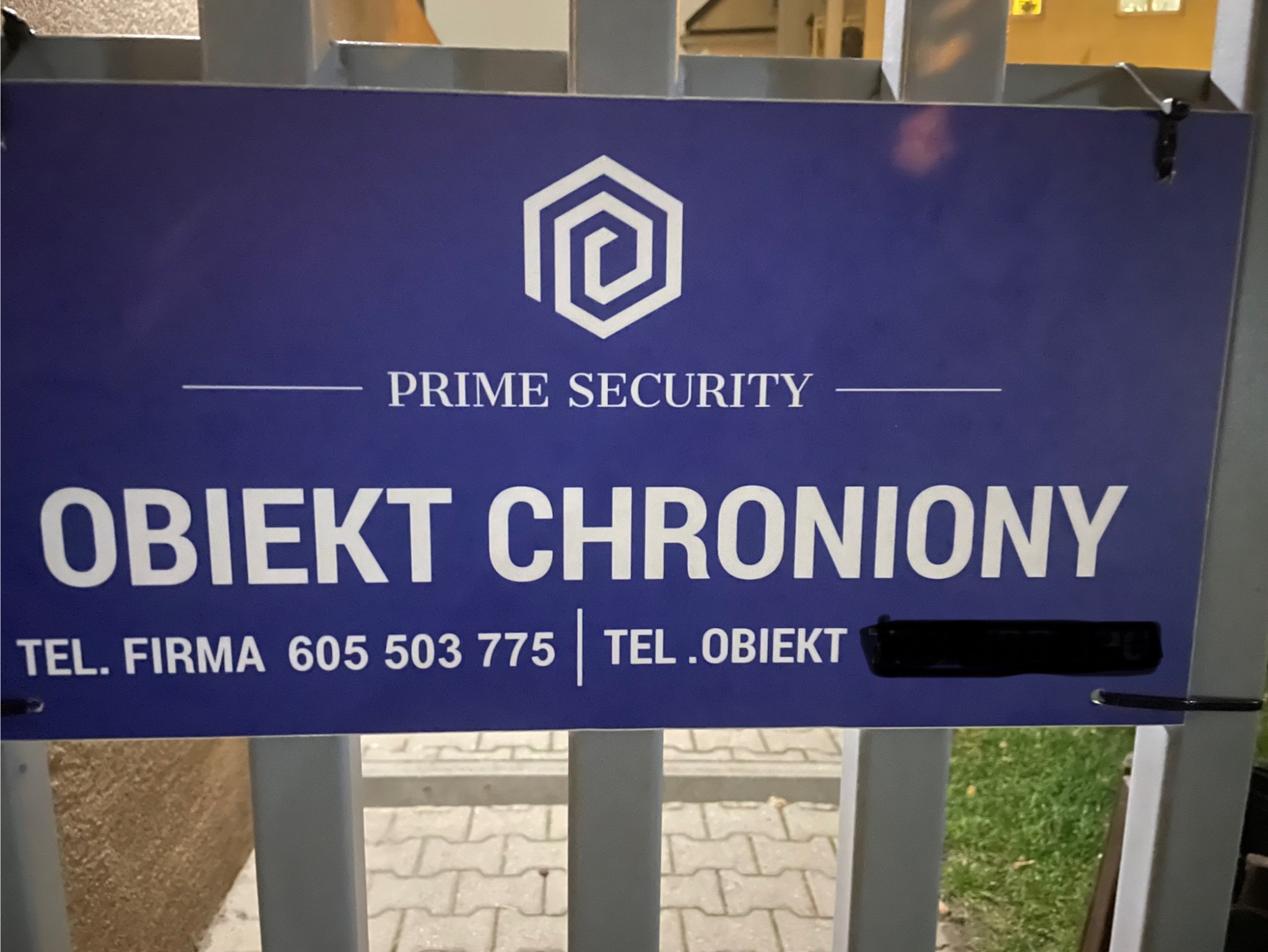 Niebieska tablica informacyjna z logo i napisem 'Obiekt Chroniony' umieszczona na metalowym ogrodzeniu, z numerem telefonu firmy i zasłoniętym numerem telefonu obiektu.