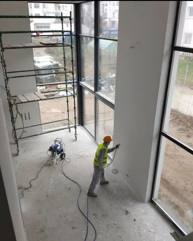 Pracownik w kasku i kamizelce odblaskowej, używając malarskiego pistoletu natryskowego, maluje wnętrze wysokiego pomieszczenia z dużymi oknami, widoczne rusztowanie i urządzenie do malowania...