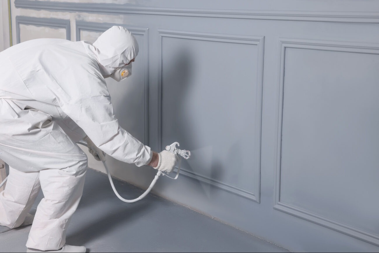 Pracownik w kombinezonie ochronnym maluje natryskowo szarą ścianę z dekoracyjnymi panelami.