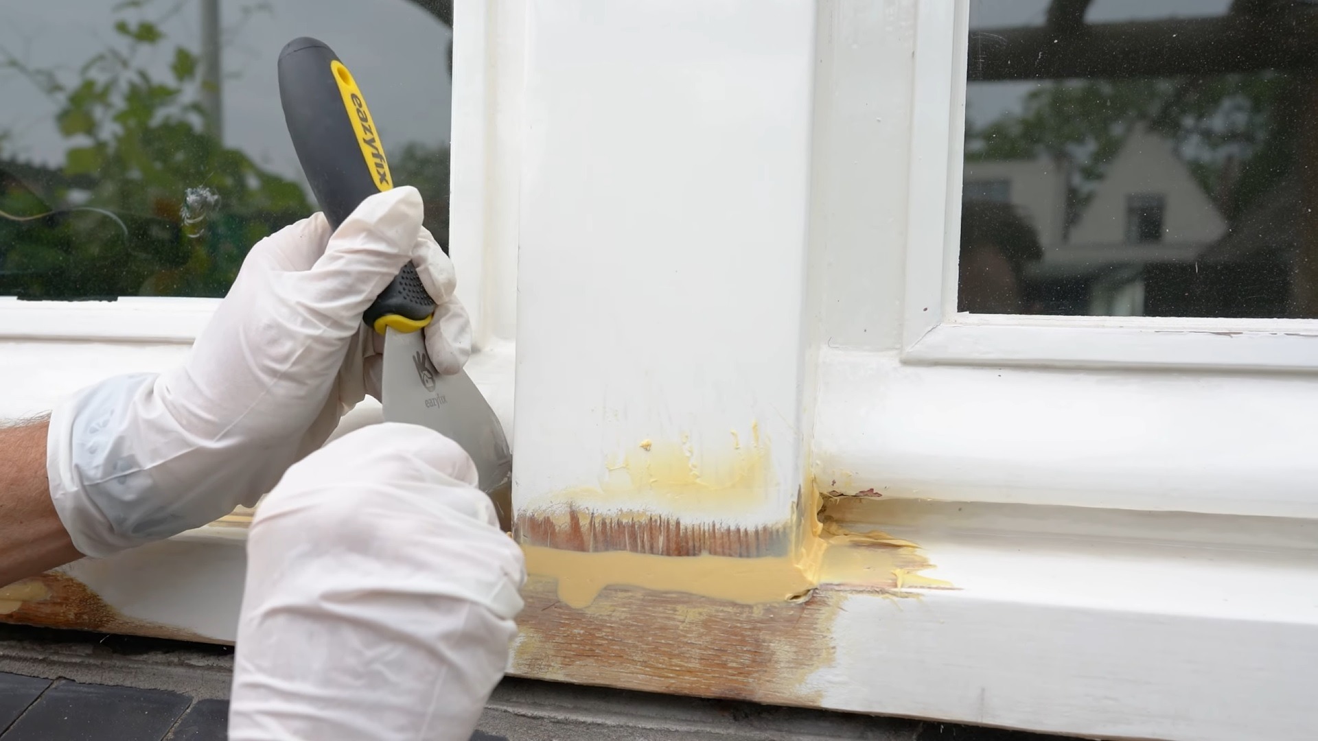 Renowacja drewnianej ramy okiennej: pracownik w białych rękawicach szpachlą Easyfix usuwa starą farbę i wypełnia ubytki szpachlą w kolorze kremowym, przygotowując powierzchnię do malowania.
