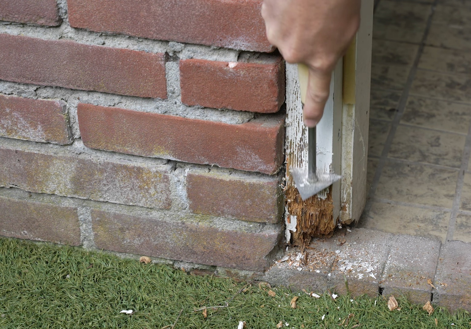 Przygotowanie do malowania elewacji: usuwanie łuszczącej się farby z drewnianego elementu przy ceglanej ścianie za pomocą szpachelki.
