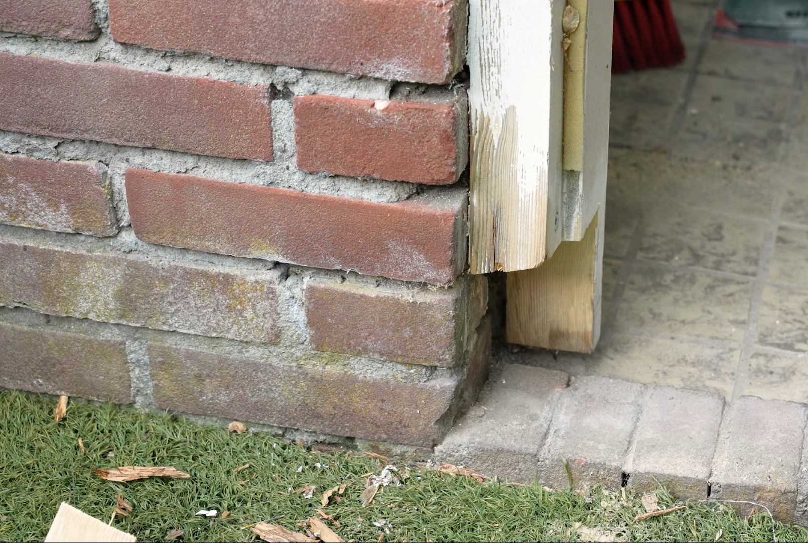 Fragment ceglanej elewacji z drewnianą, niedomalowaną ramą okienną; widoczne ślady wcześniejszych warstw farby i odłupane fragmenty drewna.