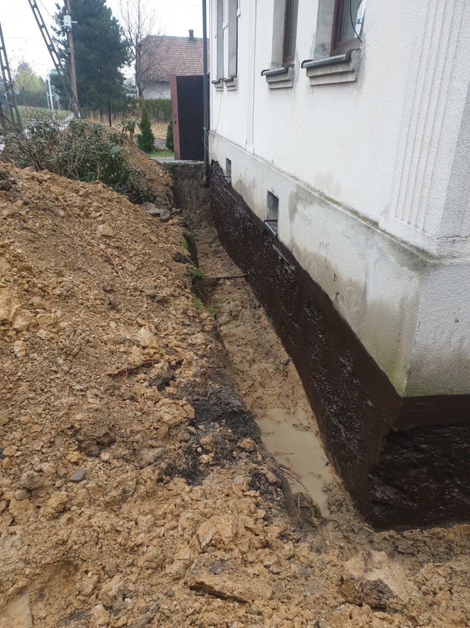 Fundament budynku po zabezpieczeniu hydroizolacją, widoczny wykop z ziemią i częściowo zakryte okienka piwniczne. Błoto w wykopie.