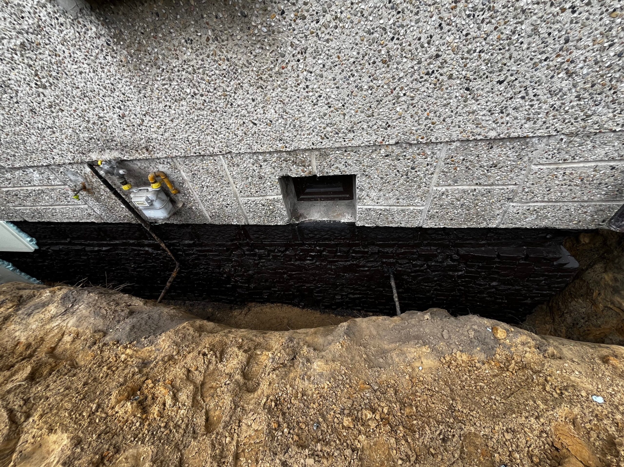 Fundament po zabezpieczeniu czarną substancją izolacyjną, widoczny wykop ziemny i fragment ściany budynku z licznikiem gazu.