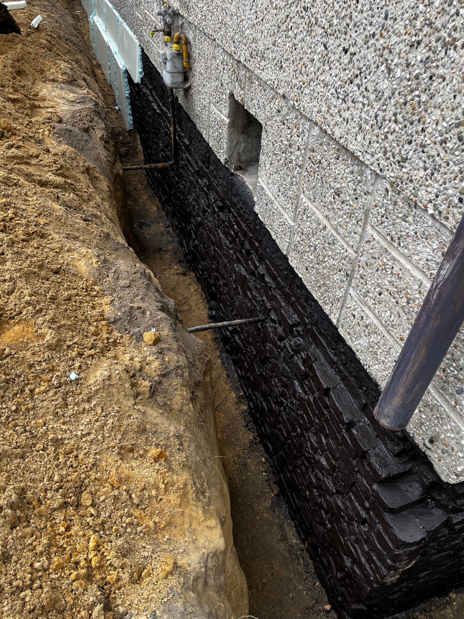 Zabezpieczony fundament budynku z izolacją termiczną i hydroizolacją, widoczny wykop z ziemią. Ściana budynku wykończona drobnym kamieniem.