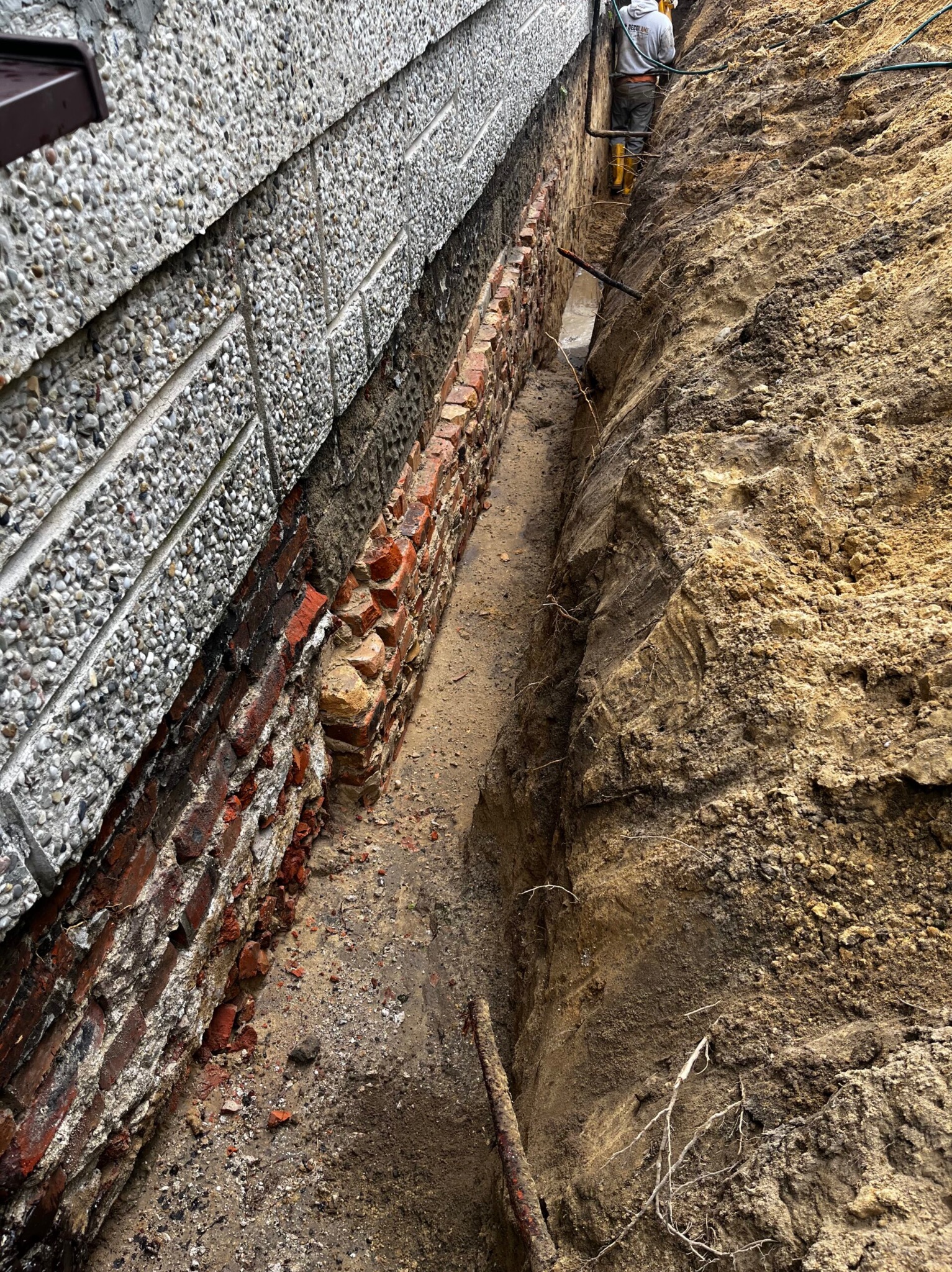 Odkopane fundamenty z cegły i betonowej ściany, widoczne wykopaliska ziemne. Osoba w oddali. Prace przygotowawcze do zabezpieczenia fundamentów.