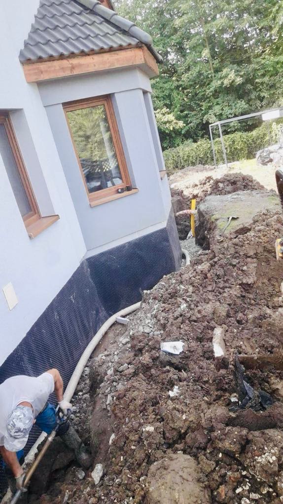 Prace ziemne przy budynku z izolacją fundamentów: drenaż, folia kubełkowa. Pracownik kopie rów.