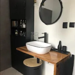 Nowoczesna łazienka z białą umywalką nablatową z czarnym obramowaniem, drewnianym blatem i okrągłym lustrem na skórzanym pasku, czarna szafka z otwartymi półkami na kosmetyki.